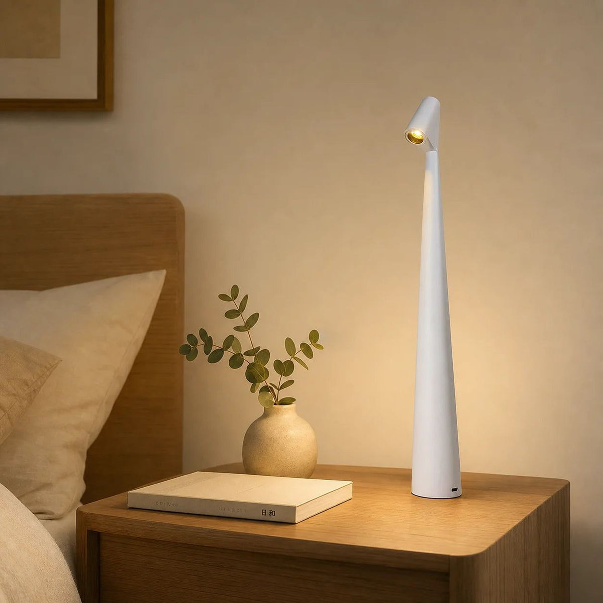 Lampe de bureau design Ambra blanche posée sur une table de chevet, ambiance soir