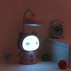 Lampe de bureau enfant Câlin rose en veilleuse dans la pénombre, visage lumineux, avec un réveil et un petit cactus à côté