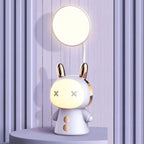 Lampe de bureau enfant Câlin violet, lumière du haut allumée, sur un socle rond devant un mur à lattes violet pâle