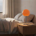 Lampe de bureau design Stellaria, globe en verre orange éteint sur une table de chevet en bois clair, en lumière du jour