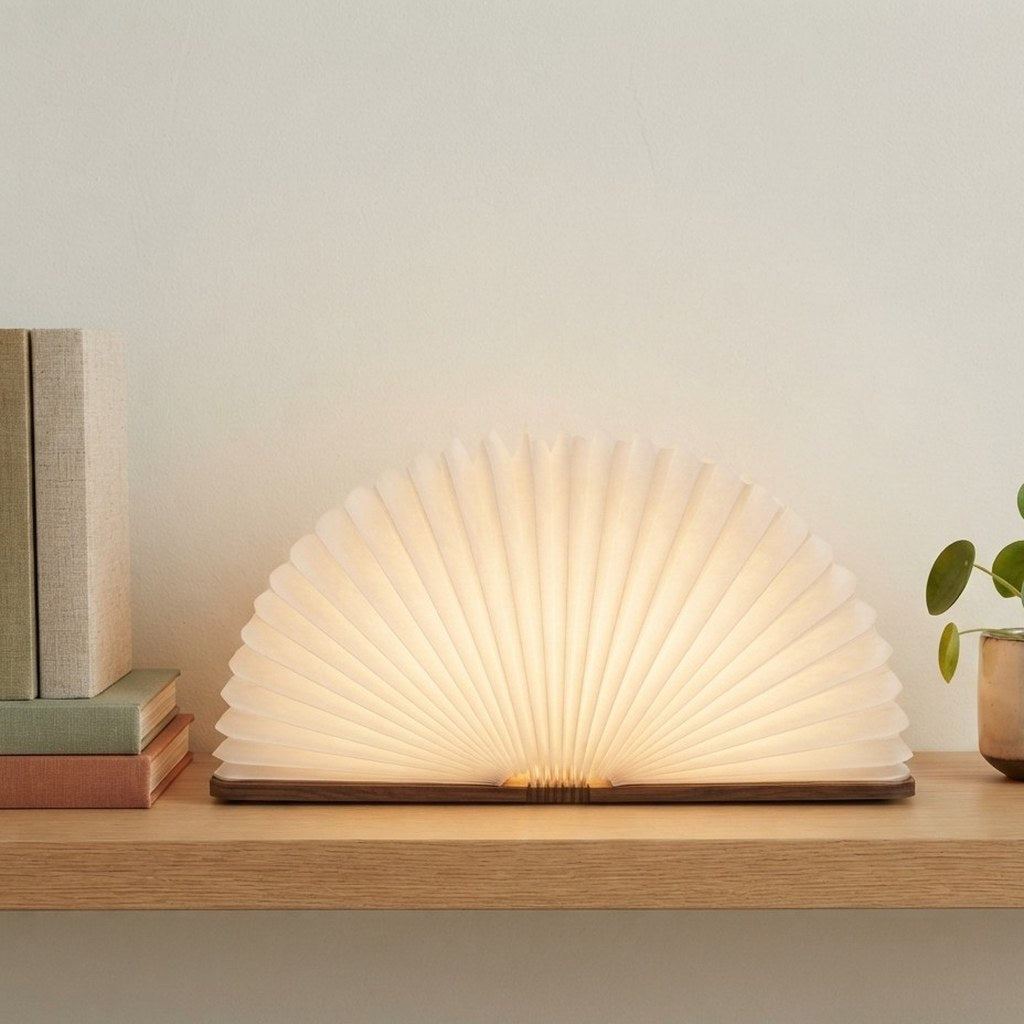 Lampe livre Conte ouverte sur une étagère en chêne clair avec des livres et une plante, en journée — Candeora